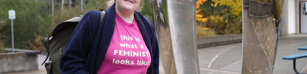a student wearing a pink shirt reading "This is what a feminist looks like" and a navy coat smiling proudly by a silver statue on campus