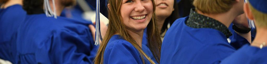 a row of graduating student with their backs turned to the camera except one young woman who turns around smiles to her family