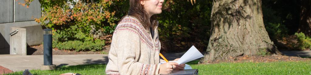 a young woman in a sweater sits and studies on blanket on the grass outside the theatre building on campus on a sunny fall day