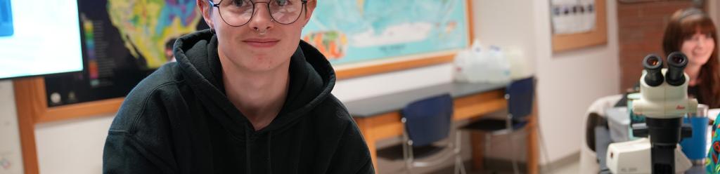 A science student with short dyed green hair and eyebrow piercing in a classroom with a large microscope