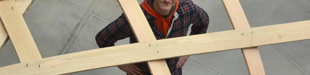 A 3D arts student, a young man wearing flannel, proudly posing underneath a wooden sculpture he made