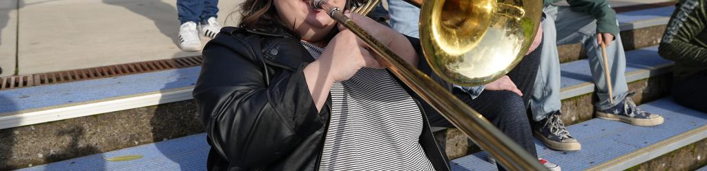 a heavy-set young woman with a trendy leather jacket and bangs joyfully plays the trombone while her fellow music students laugh happily