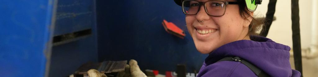 a welding student, a young Latina woman in a purple sweatshirt, smiling at a welding station