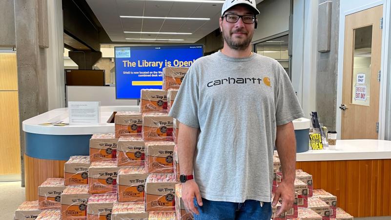 Chad Drake poses stands in front of the mountain of ramen noodle display that illustrates how much a student can save by choosing classes that use OER for class materials.