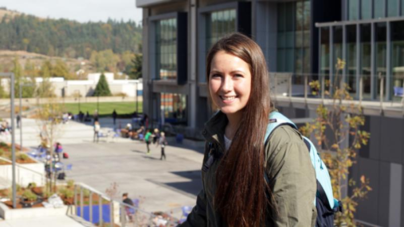 A young woman with a backpack smiling in front of Center Building on main campus