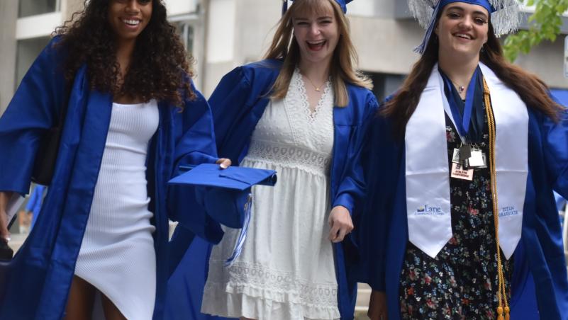 Three Lane Community College graduates in blue gowns walk together smiling, wearing dresses and caps, with one in honors regalia.