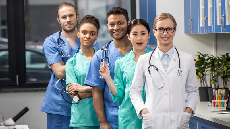 Group of diverse medical students or young healthcare professionals in scrubs and a lab coat, smiling in a clinical setting.
