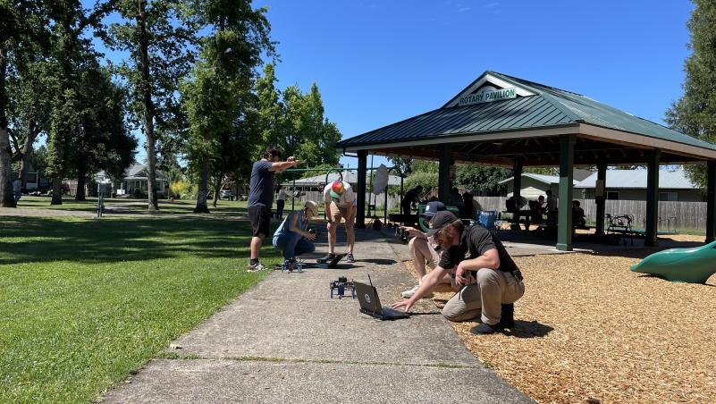 Drone students work to survey trees in a sunny park with trees and a pagoda  in the background