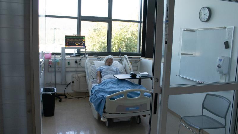 Simulation hospital room with a mannequin in bed, medical monitor, and equipment by the window.