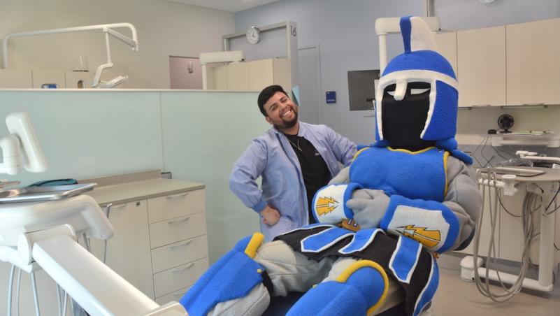 Ty the Titan sitting in an exam chair in the new dental clinic next to a smiling dental hygiene student, a young man of color