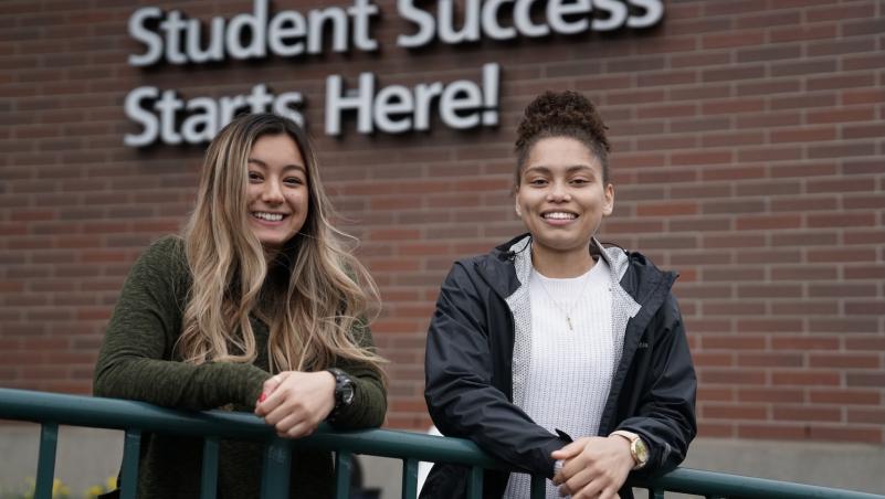 two students, smiling young women, pose in front of the "student success starts here" sign on Building 1