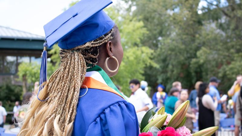 a black woman, an international student, in blue cap and gown turned from the camera, holding flowers and looking over the crowd at Commencement