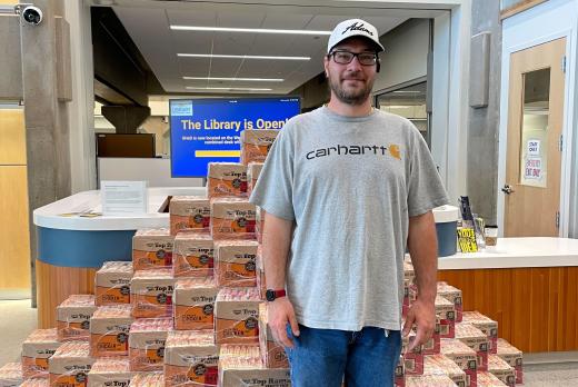 Chad Drake poses stands in front of the mountain of ramen noodle display that illustrates how much a student can save by choosing classes that use OER for class materials. 