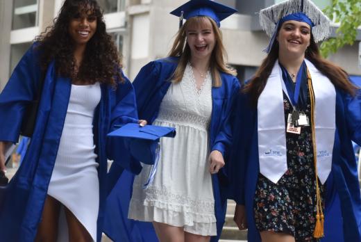 Three Lane Community College graduates in blue gowns walk together smiling, wearing dresses and caps, with one in honors regalia.