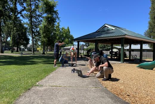 Drone students work to survey trees in a sunny park with trees and a pagoda  in the background