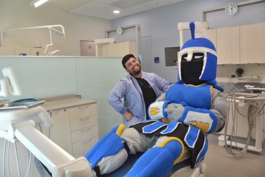 Ty the Titan sitting in an exam chair in the new dental clinic next to a smiling dental hygiene student, a young man of color