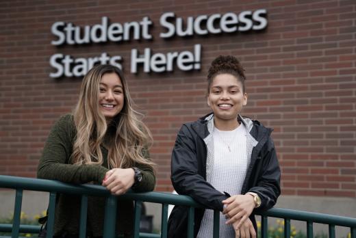 two students, smiling young women, pose in front of the "student success starts here" sign on Building 1