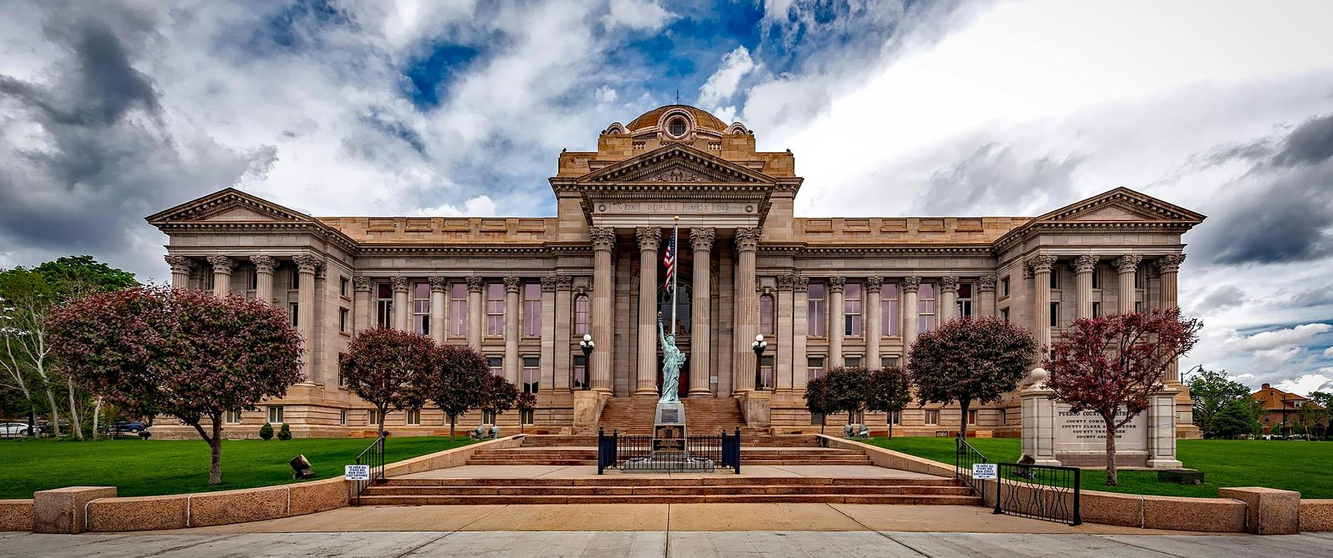 a courthouse in neoclassical style with a liberty statue in front and a dramatic blue sky behind