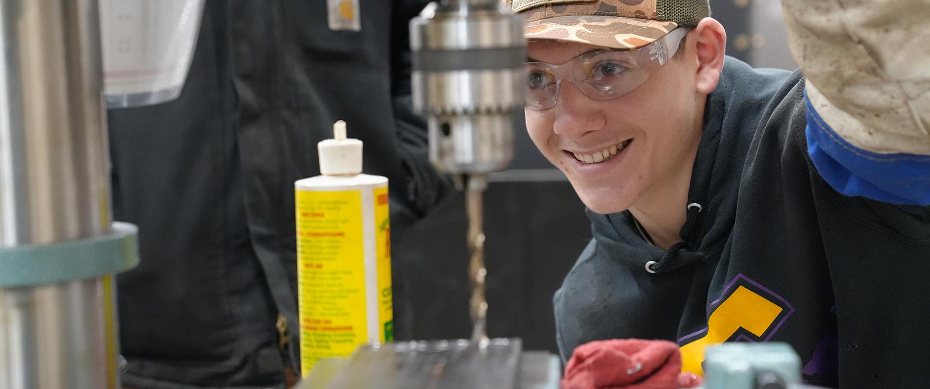 a millwright apprentice in a camo hat smiles and uses a drill press