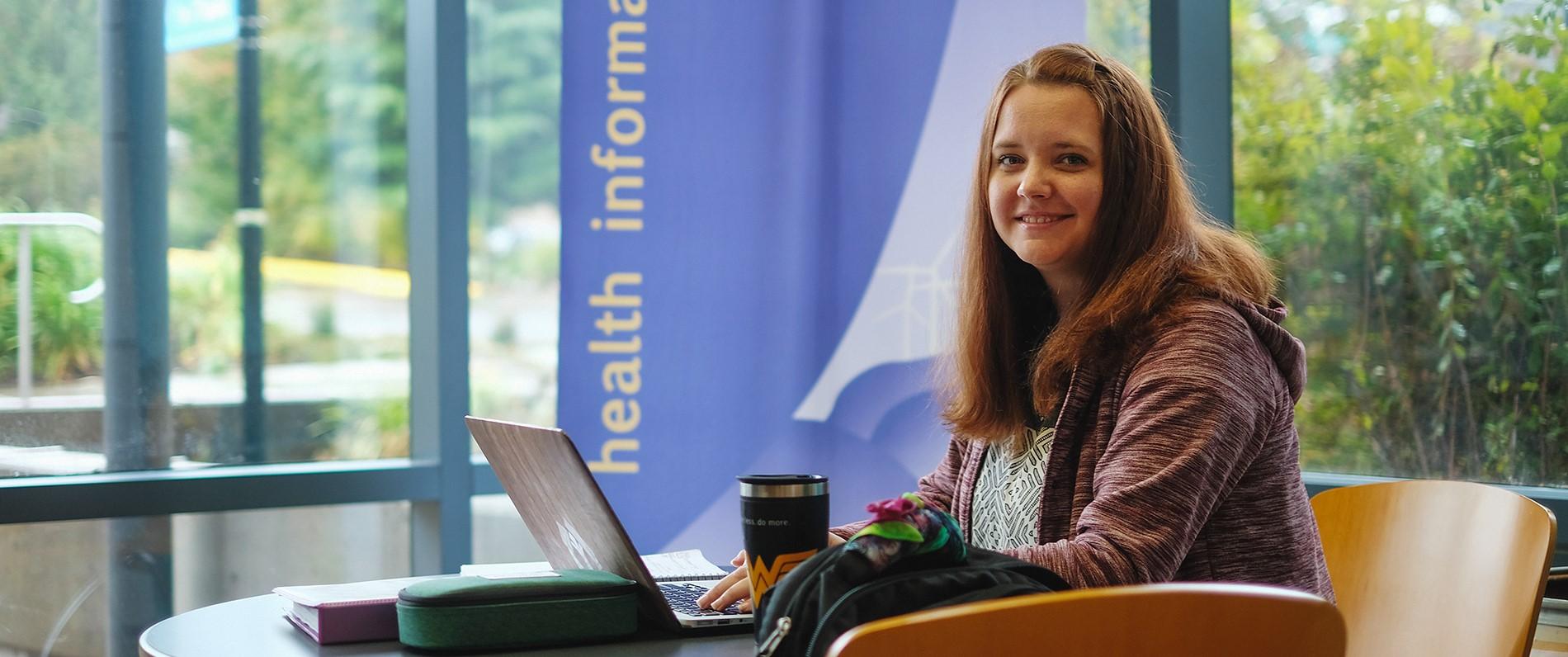 Pre-professional health professions student with a laptop smiles in a sunny lobby
