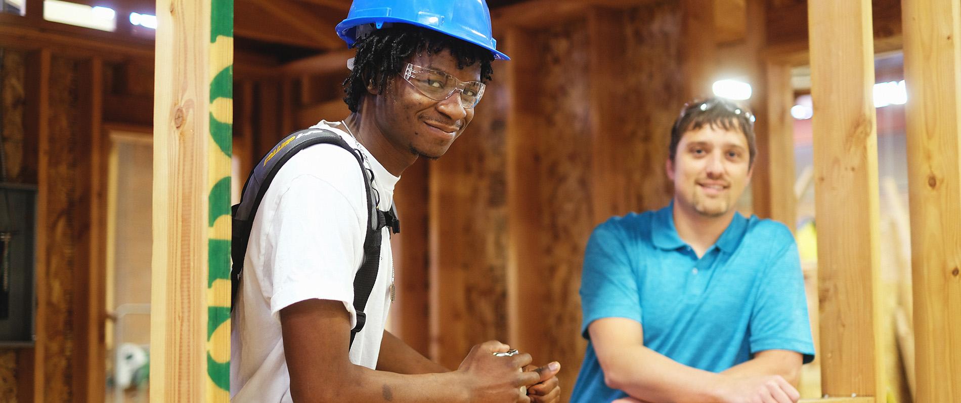 two construction technology students, one wearing PPE, pose and smile in the frame of a wall they're building