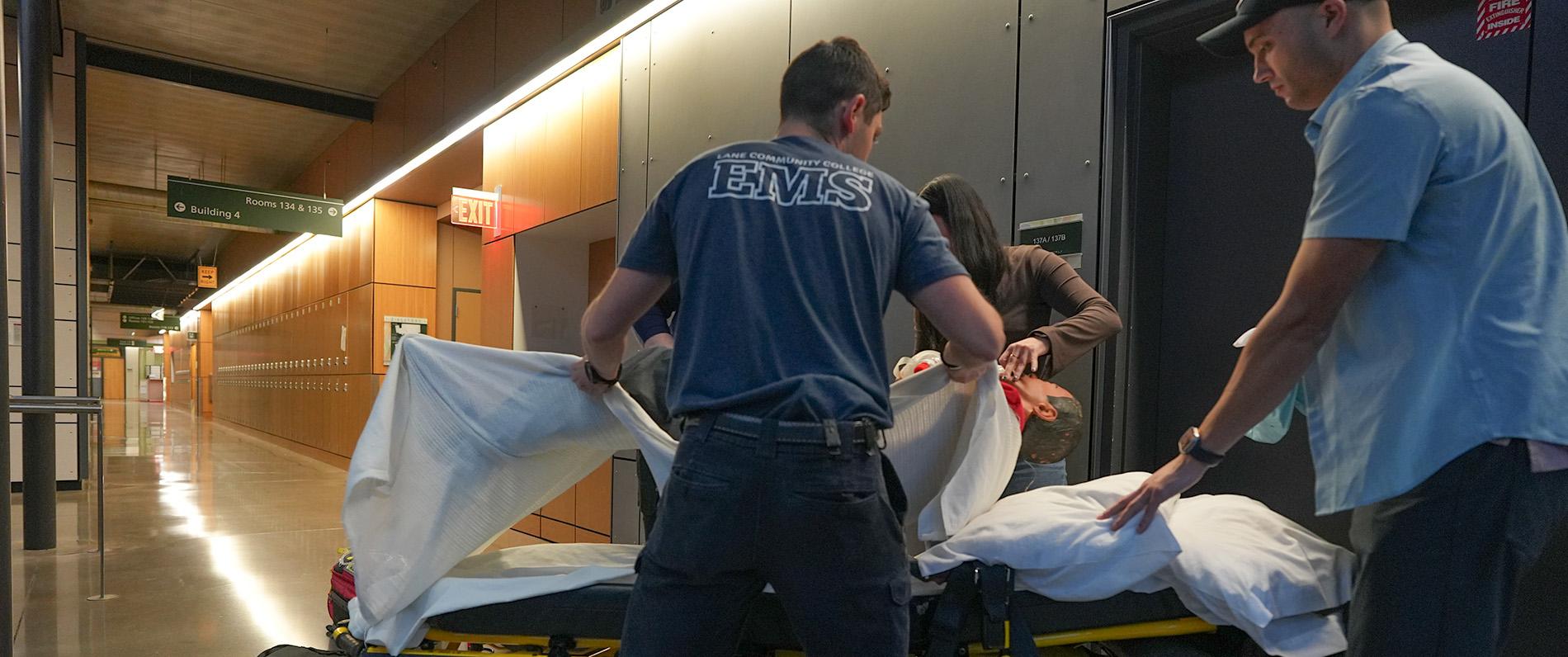two EMT/Paramedicine students practice moving a dummy onto a gurney in the Health Professions building