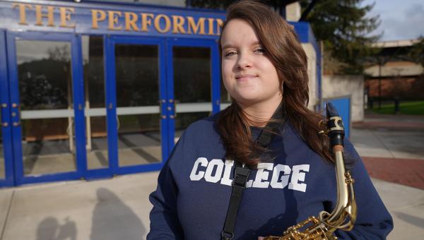 a music student, a young woman in hoodie reading "College," poses proudly with her saxophone in front of the music building