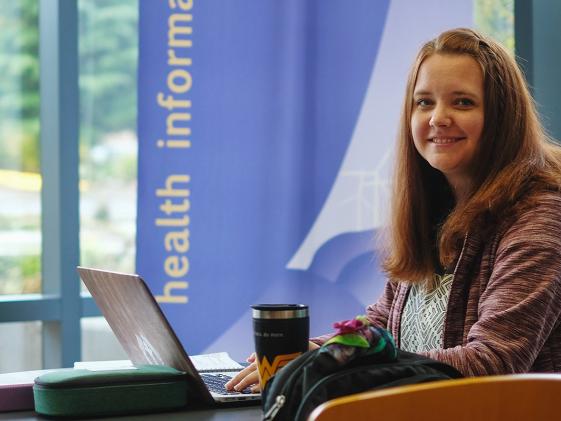 Pre-professional health professions student with a laptop smiles in a sunny lobby