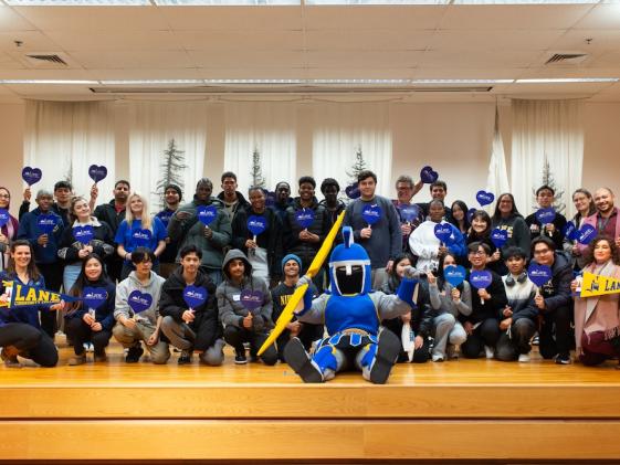 a large group of international students and staff smiling, holding LCC signs, and posing with mascot
