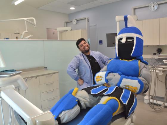 Ty the Titan sitting in an exam chair in the new dental clinic next to a smiling dental hygiene student, a young man of color