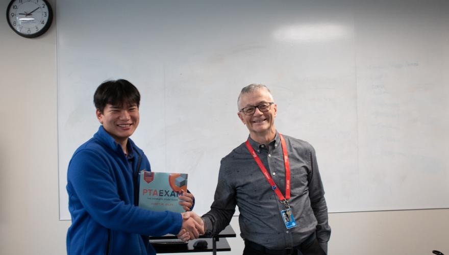 Physical Therapist Assistant instructor Marc Duyck gives a text book to a smiling student, Ramsan Aung, a young Asian man