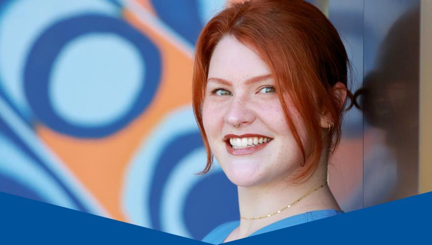 Abby, red haired person wearing blue scrubs leaning against a mural of abstract artwork.