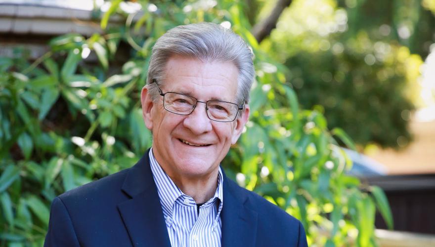 Alan Yordy, a mature individual with a suite, no tie, standing with hads linked in from smiling to the camera in a garden.