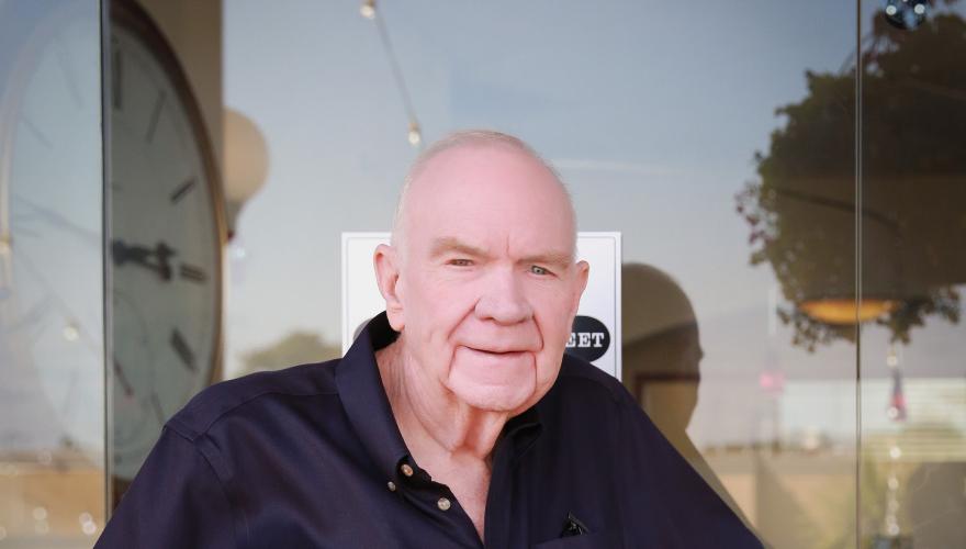 A mature individual with a black button up shirt grasping a handle of a glass door smiling at the camera