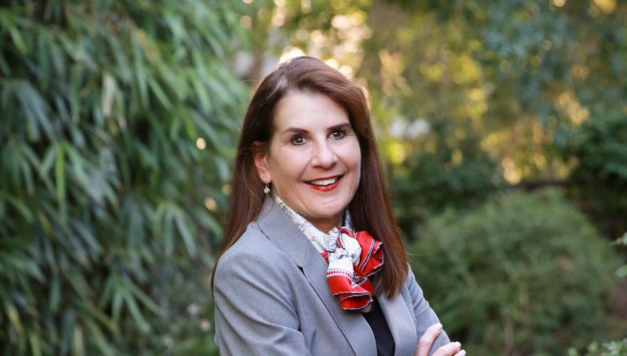 Irene, a mature individual with a grey suit and a red and white ascot smiling at the camera with arms crossed. 