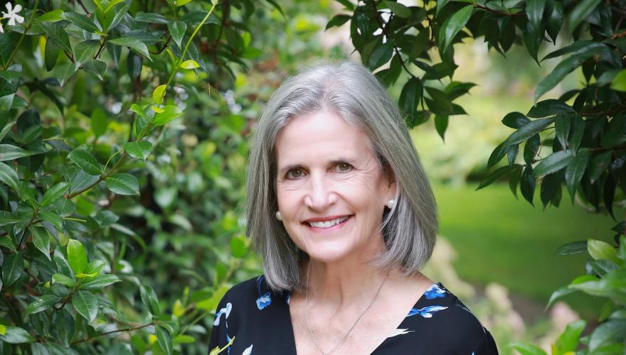 Jenny, a matuure individual with a black floral shirt and jeans smiling into the camera while surrounded by trees. 