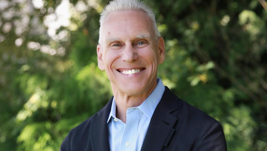 Steve, a mature individual with a black suit and blue shirt smiling at the camera with arms across chest in front of a cedar tree. 