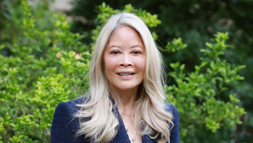 Paula, a mature inividual with long blond hair down on her shoulders wearing a suit with gold buttons in a garden. 
