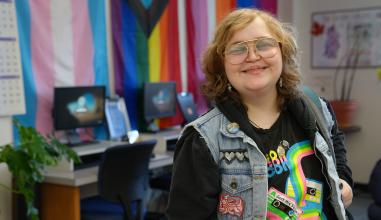A student with a warm smile, big glasses, and a jean jacket with pins in the Gender Equity Center with pride flags in background