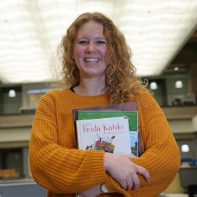 a smiling young woman in a big yellow sweater holding textbooks in the library