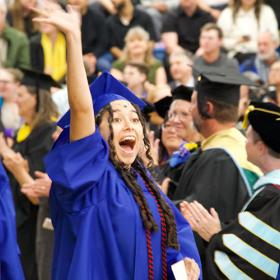 a graduating student in blue cap and gown excitedly waves to her family at 2024 Commencement