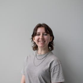 a young woman with a gray t-shirt, dark hair, and black eyeliner smiling broadly