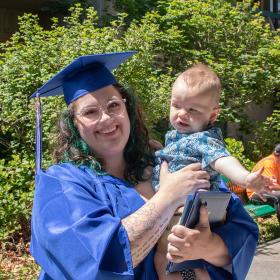 a happy woman with glasses, tattoos, dyed hair, and her blue cap and gown smiles and holds her baby at Commencement