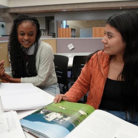 Two friends, young women in cozy winter jackets, studying in the library and smiling