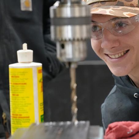 a millwright apprentice in a camo hat smiles and uses a drill press