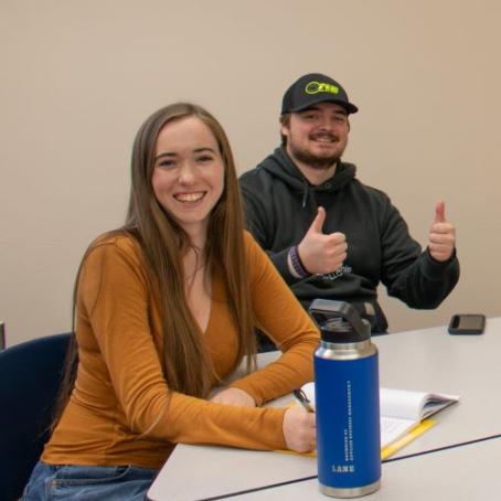 two bachelors business students, a young woman smiling, and a young man wearing a baseball cap giving a thumbs up