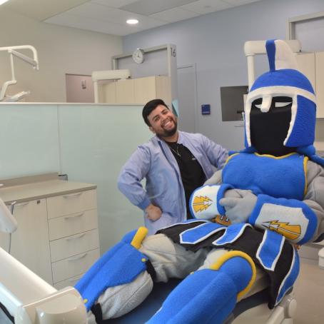 Ty the Titan sitting in an exam chair in the new dental clinic next to a smiling dental hygiene student, a young man of color