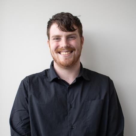 a tall young man with red hair and beard, smiling in a dark, button up shirt