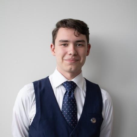 a straight-lace young man in a suit vest and tie, smiling seriously at the camera
