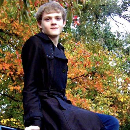 Early College and pre med student Nathan smiles while sitting on a bench with autumn foliage in background